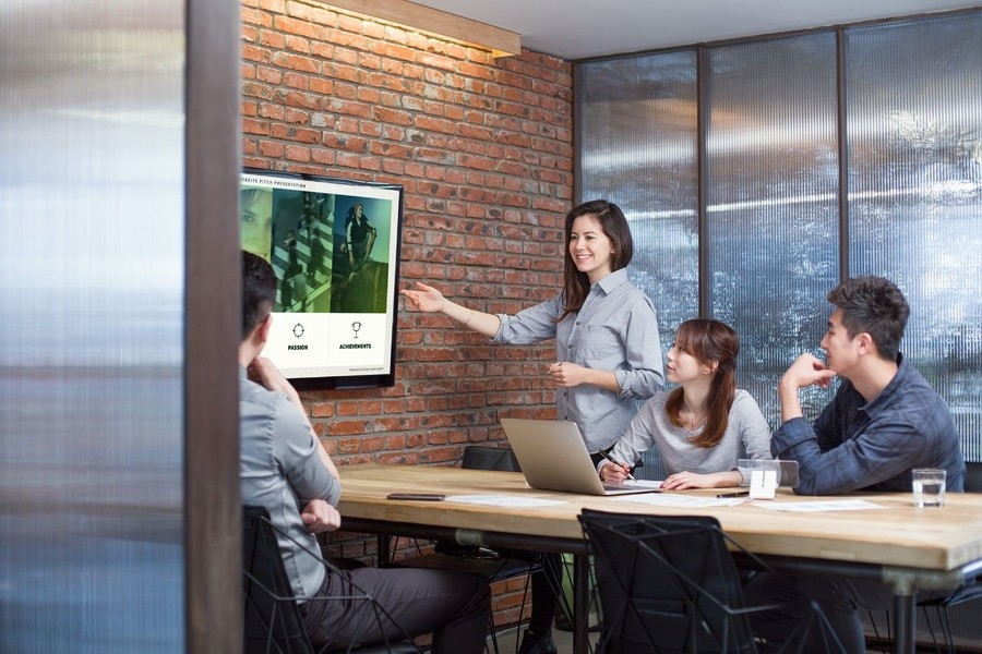 Meeting room with one person presenting on a screen in a brick-walled room with glass partitions.